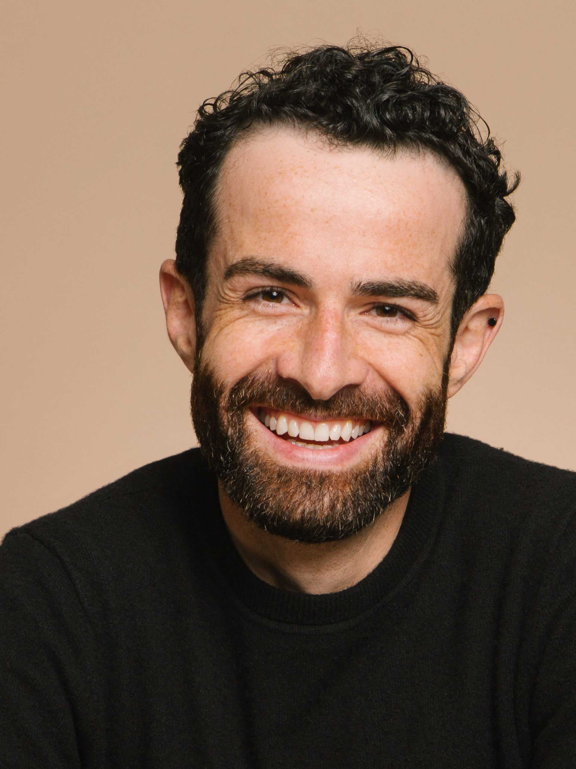 A portrait of a cheerful man with curly hair and beard, smiling widely, showing white teeth. He has dark, short, curly hair, and a groomed beard. Freckles are visible on his face and he's wearing a black crew neck sweater. The background is a warm, neutral tone.